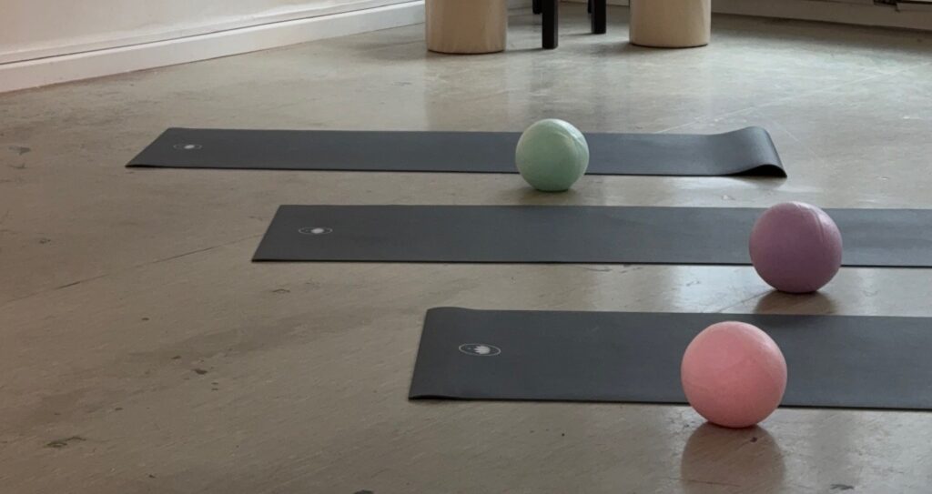 Drei Pilatesbälle in Grün, Lila und Rosa liegen auf ausgelegten Matten in einem Studio.