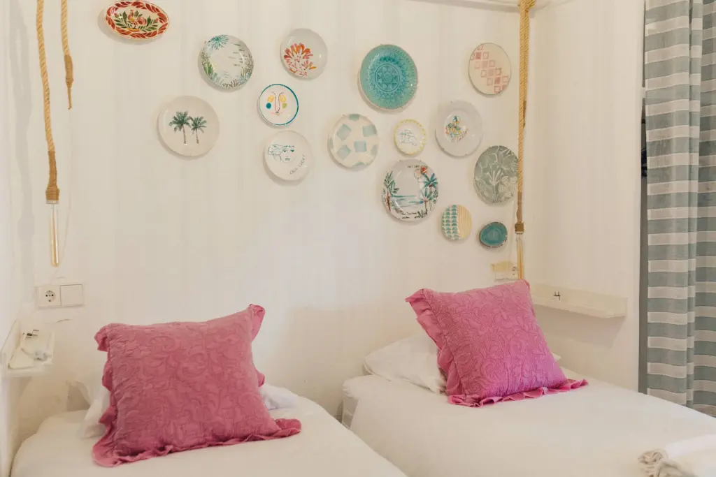 Zimmer mit zwei Einzelbetten und rosa Kissen, an der Wand eine Sammlung dekorativer Teller.