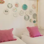 Zimmer mit zwei Einzelbetten und rosa Kissen, an der Wand eine Sammlung dekorativer Teller.