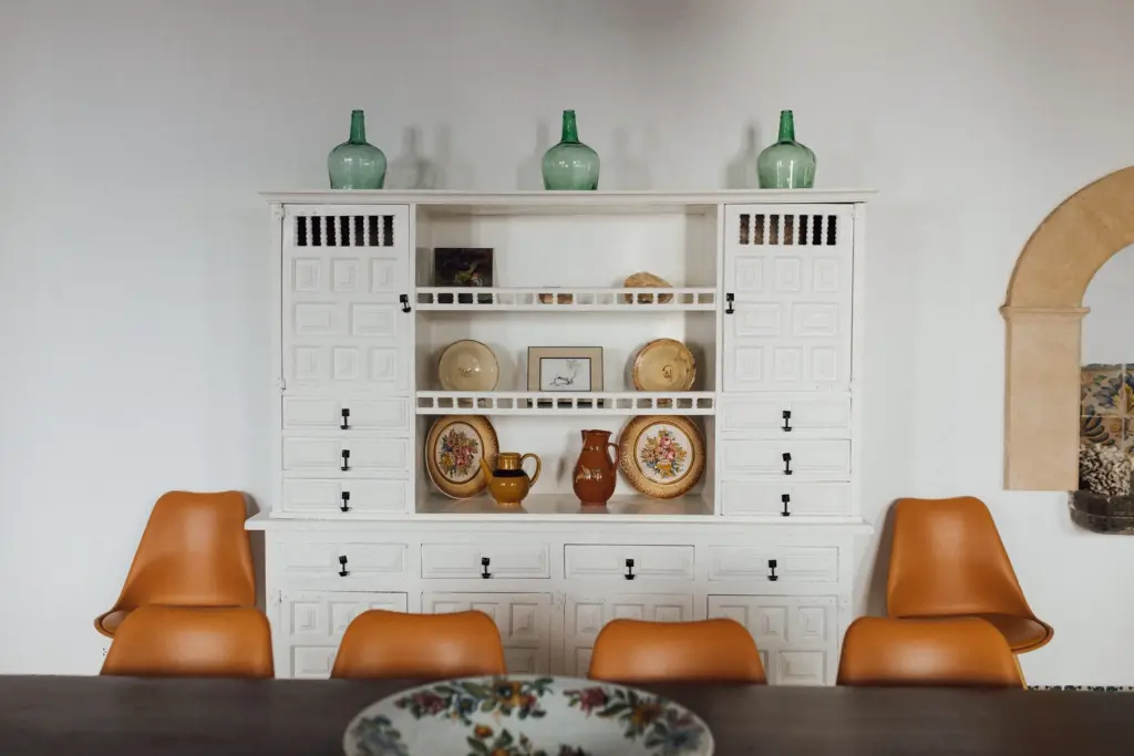 Weiße Vitrine mit mallorquinischer Keramik, davor zwei orangefarbene Lederstühle.
