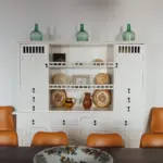 Weiße Vitrine mit mallorquinischer Keramik, davor zwei orangefarbene Lederstühle.
