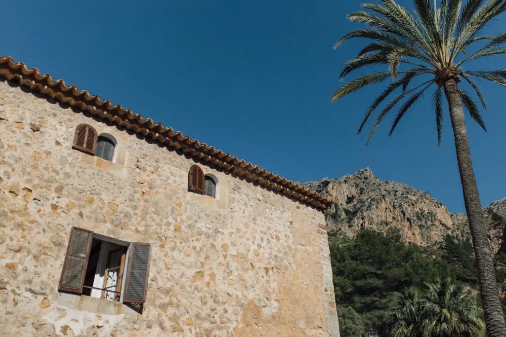 Mallorquinische Finca aus Naturstein mit Ziegeldach, davor eine Palme vor Felsen und blauem Himmel.