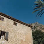 Mallorquinische Finca aus Naturstein mit Ziegeldach, davor eine Palme vor Felsen und blauem Himmel.