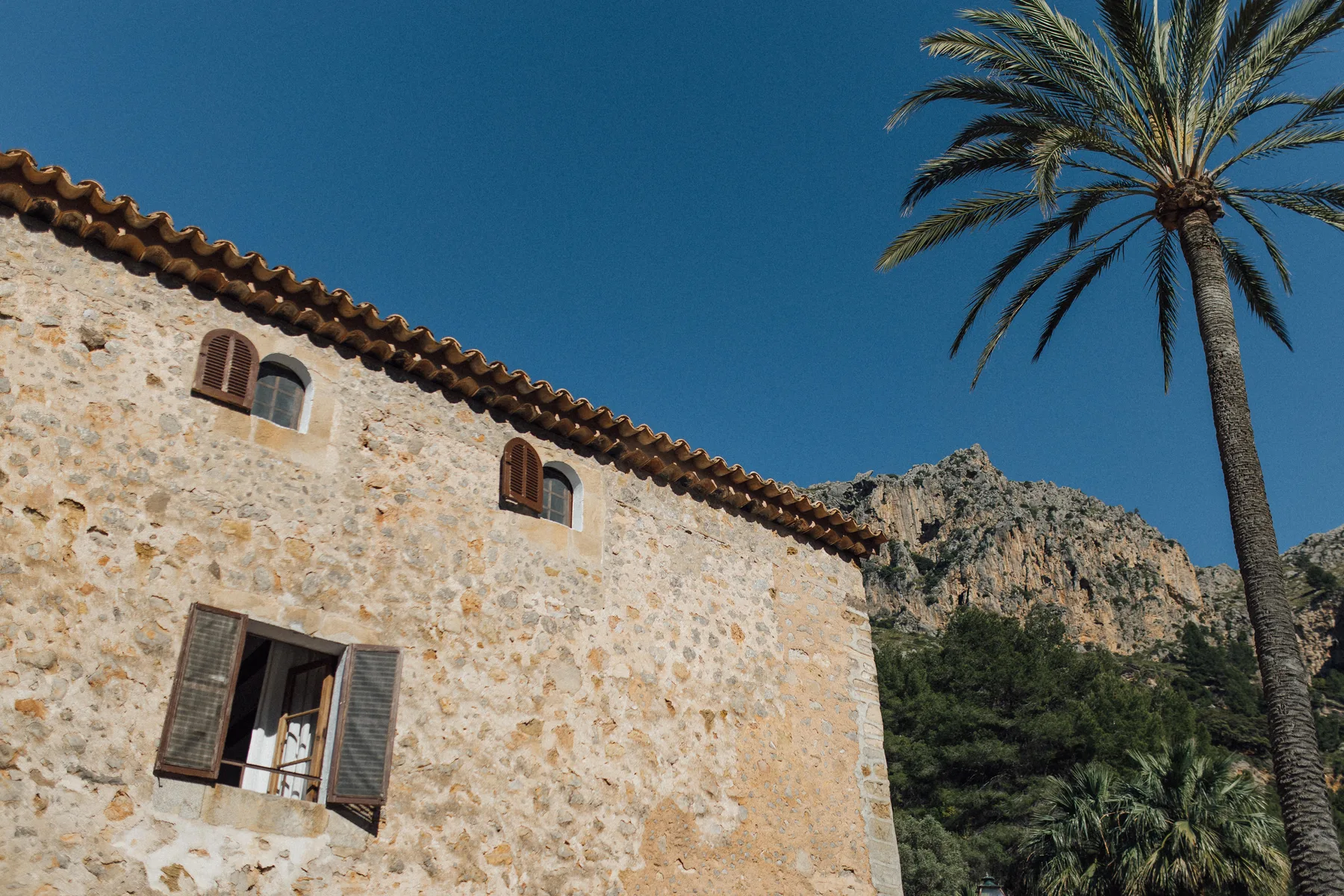 Mallorquinische Finca aus Naturstein mit Ziegeldach, davor eine Palme vor Felsen und blauem Himmel.