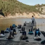 Yogagruppe liegt zur Schlussentspannung auf Matten auf einem Holzsteg am Wasser, rechts steht die Kursleiterin.