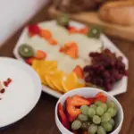 Frühstücksplatte mit Obst und einer Schale Trauben und Erdbeeren, dahinter unscharf ein Brotkorb.