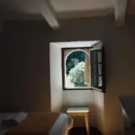Zimmer mit Bogenfenster, auf dem dunklen Holzboden liegt ein Lichtfleck; das Bett trägt eine ockerfarbene Decke.