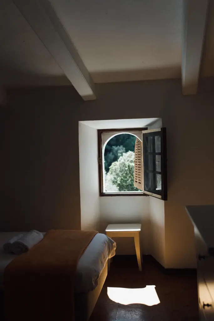 Zimmer mit Bogenfenster, auf dem dunklen Holzboden liegt ein Lichtfleck; das Bett trägt eine ockerfarbene Decke.