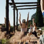 Yogagruppe unter einer Pergola aus Schilf, in der Bildmitte streckt eine Frau von hinten gesehen beide Arme nach oben; im Vordergrund unscharfe Blüten.