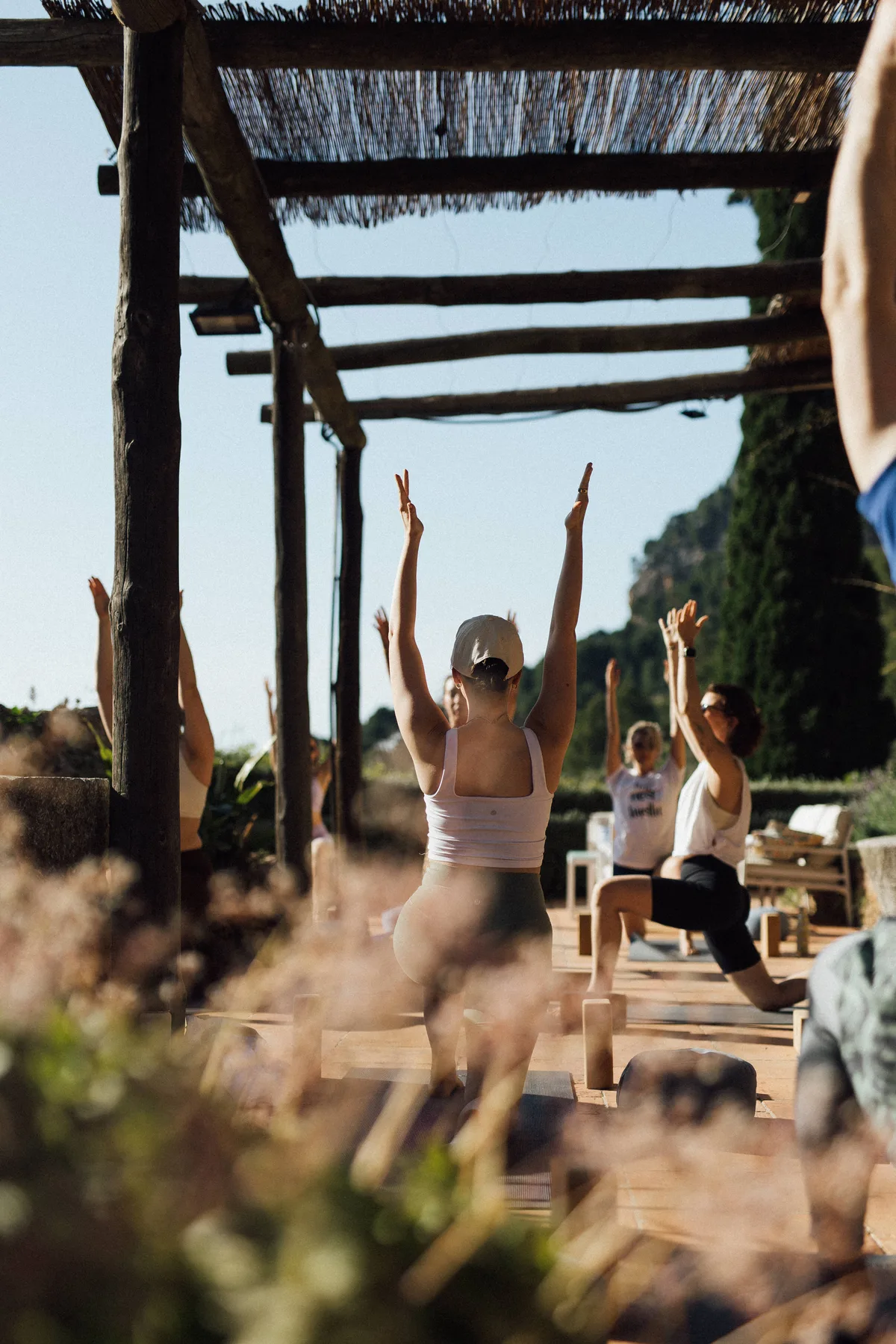 Yogagruppe unter einer Pergola aus Schilf, in der Bildmitte streckt eine Frau von hinten gesehen beide Arme nach oben; im Vordergrund unscharfe Blüten.