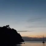 Bucht in der blauen Stunde, das Kap als dunkle Silhouette, rechts ein Segelboot, über dem Horizont ein oranger Streifen.