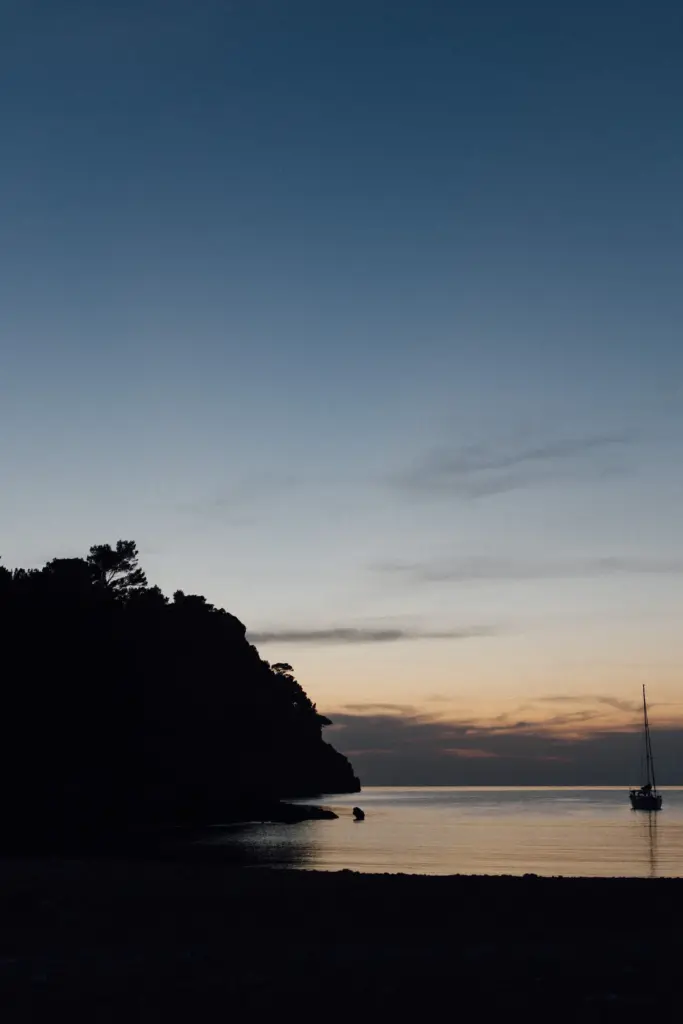 Bucht in der blauen Stunde, das Kap als dunkle Silhouette, rechts ein Segelboot, über dem Horizont ein oranger Streifen.