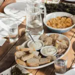 Gedeckter Holztisch mit Brotkorb, zwei Schälchen Aioli, einer Schüssel Kartoffeln, gebratenen Pimientos und Wassergläsern.