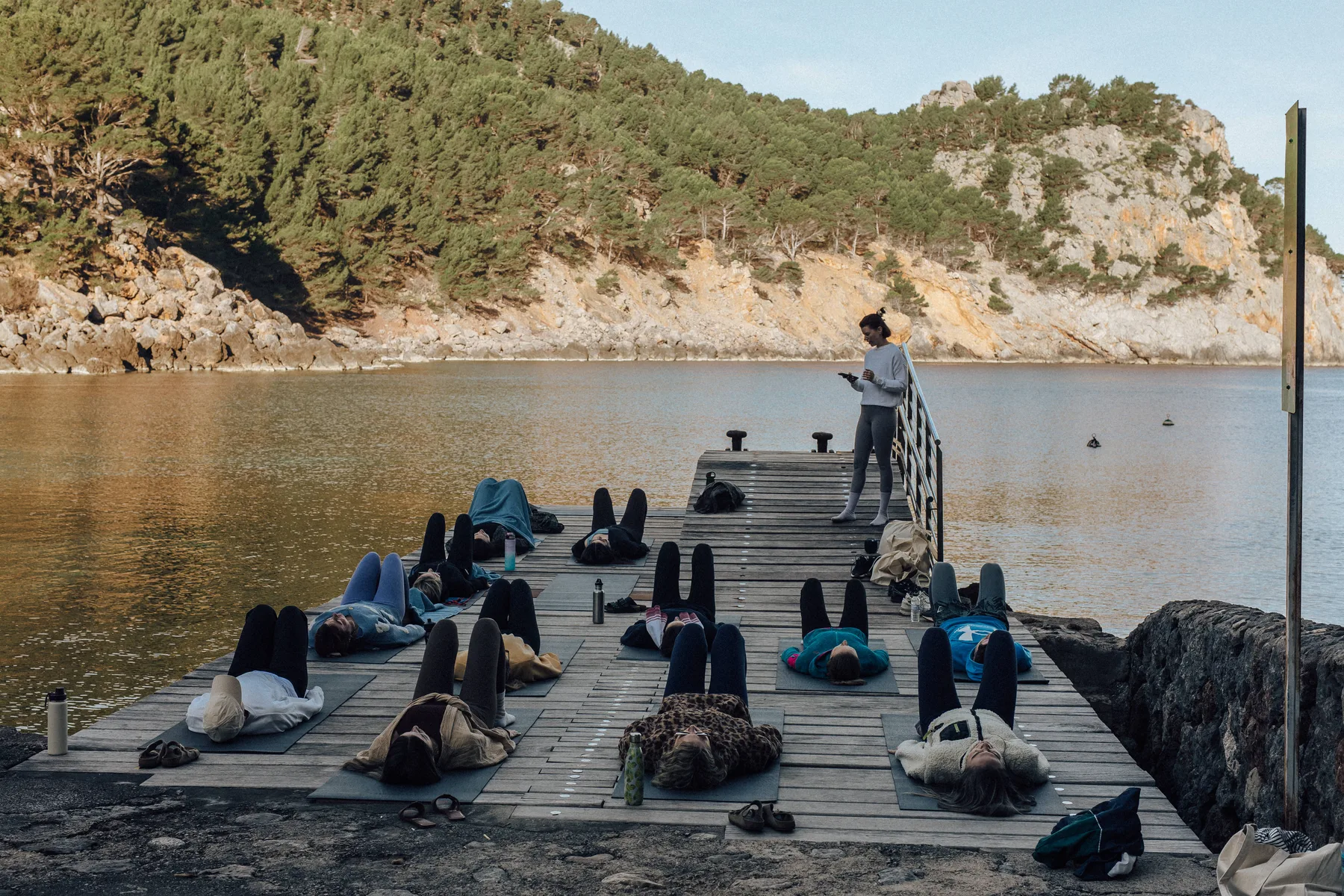Yogagruppe liegt zur Schlussentspannung auf Matten auf einem Holzsteg am Wasser, rechts steht die Kursleiterin.